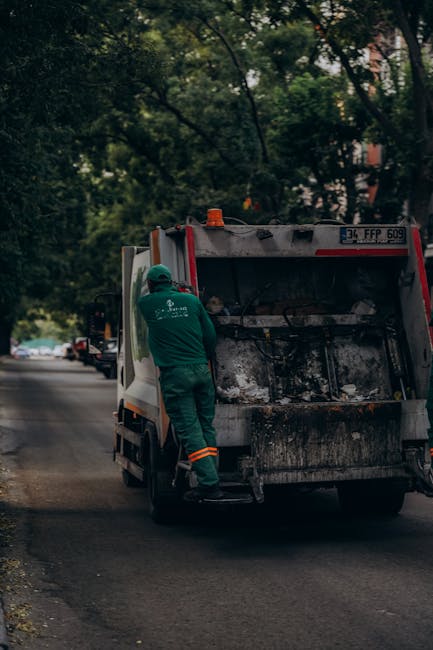 A male sanitation worker dressed in a high-visibility yellow and red safety vest and dark trousers is operating a large red refuse collection vehicle on a street. The vehicle, positioned with its rear